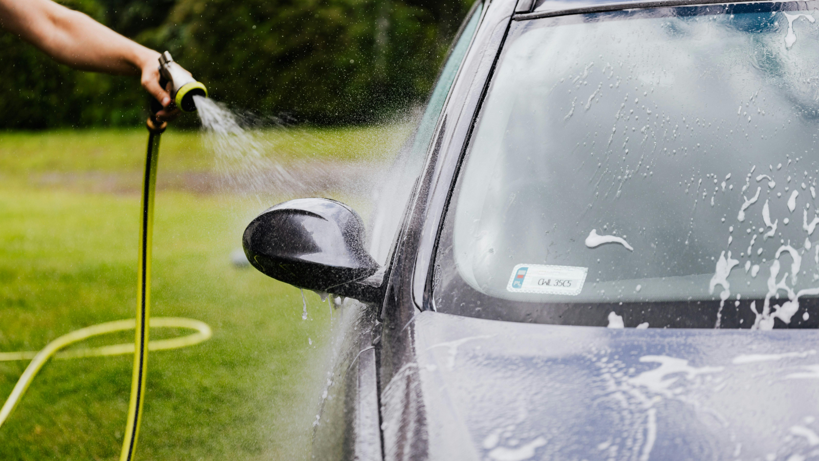Person washing a black car with a hose outdoors, rinsing off soap suds from the side mirror and windshield.