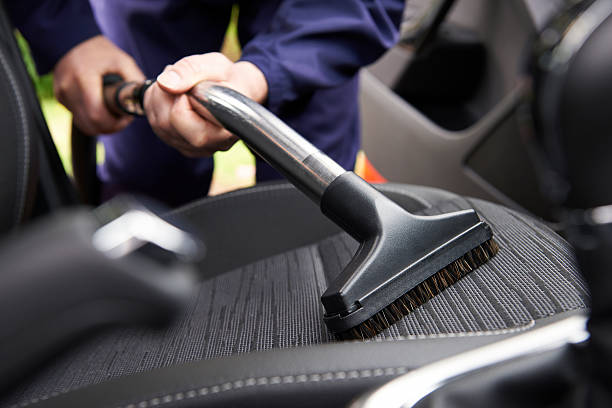 Person vacuuming a car seat with a brush attachment to remove dust, debris, and pet hair from the car interior.