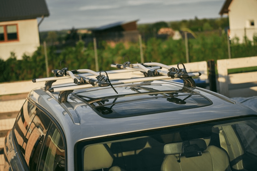 A close-up view of a silver SUV with a roof rack system designed for carrying bicycles, parked outdoors near a wooden fence, with houses and greenery in the background