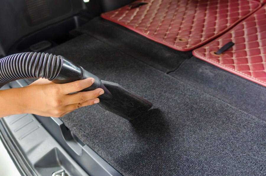 A person vacuuming the carpeted trunk area of a car, with red quilted floor mats placed on the side for cleaning