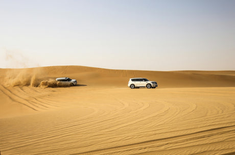 Two white 4x4 vehicles driving through sandy desert dunes, one kicking up sand while off-roading under a clear blue sky.