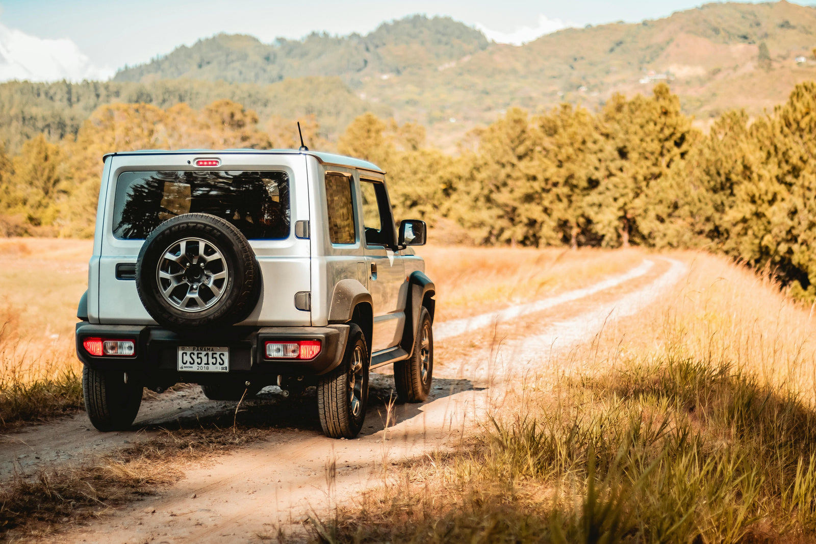 A silver off-road SUV driving along a winding dirt road surrounded by golden fields and lush green hills under a bright sky